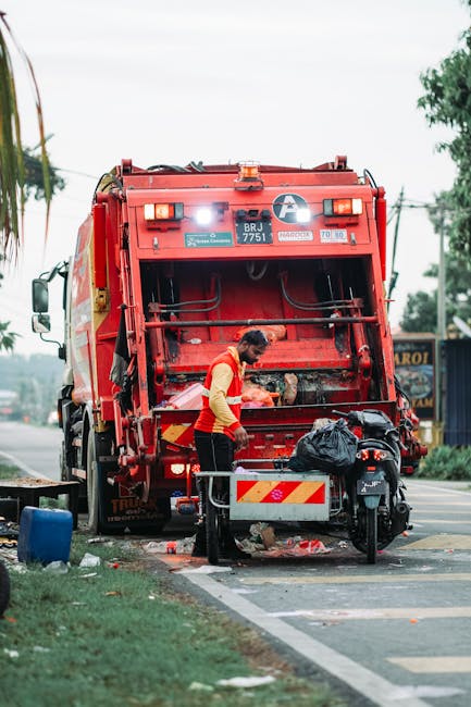 A waste collection worker in a yellow and red uniform is standing behind a large red rubbish truck, actively emptying a black refuse bag into the rear loading compartment. The truck is positioned on t