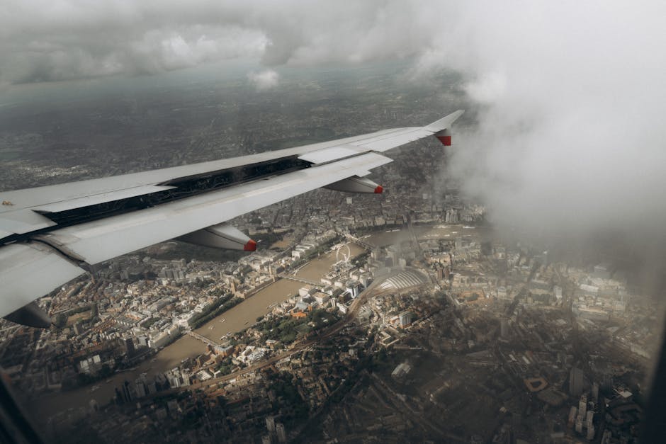 A view from an aircraft window showing the airplane's white wing with red accents extending over a sprawling urban landscape below. The wing has visible mechanical details and a smooth, metallic surfa