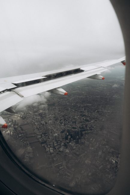 A view from an aircraft window showing the airplane's white wing with red accents extending over a sprawling urban landscape below. The wing has visible mechanical details and a smooth, metallic surfa